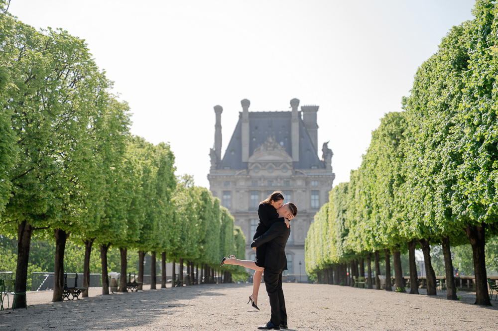 Newly engaged man lifting his fiancée for a kiss on the tree-lined allée of the Tuileries Garden in Paris, by proposal photographer Irene