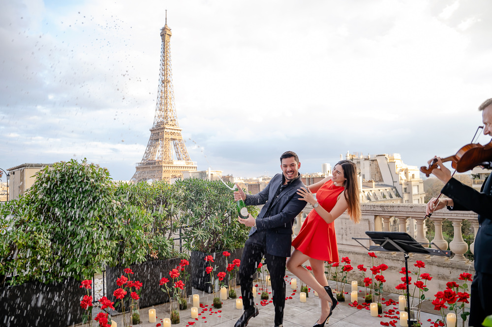 Just engaged couple popping champagne on a Paris rooftop with an Eiffel Tower view, red roses, candles and a violinist, by Irene